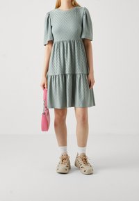 Himmelblaues, gestuftes Kleid mit Puffärmeln, das ein Chevron-Muster und kleine Punkte aufweist. Kombiniert mit beigen Clogs und einer rosa Handtasche.