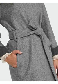 Grauer Wollmantel mit tailliertem Schnitt und Schleifendetail, ausgestattet mit dunkelgrauen Bündchen. Strukturierter Stoff mit weichem, warmem Aussehen.