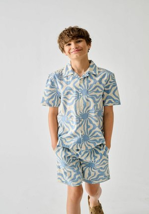 Jongen met handen in de zakken, die een bijpassend blauw en beige zonpatroon kort mouw shirt en korte broek draagt, glimlachend tegen een effen witte achtergrond.