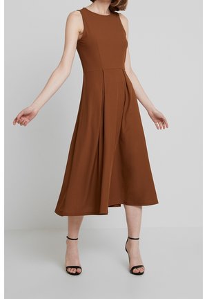 Robe midi marron sans manches avec un corsage ajusté et une jupe fluide à plis verticaux. Texture de tissu lisse, portée avec des sandales à talons noires.