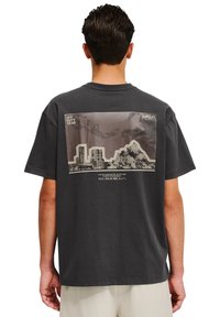 Grå t-shirt i bomull med ett stort grafiskt tryck på baksidan av en stadssiluett och berg. Korta ärmar och en avslappnad passform.