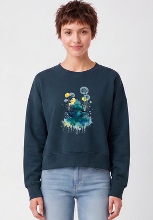 Jonge persoon met kort haar die een marineblauwe trui draagt met een kleurrijke mediterende figuur en bloemenpatroon op de voorkant.