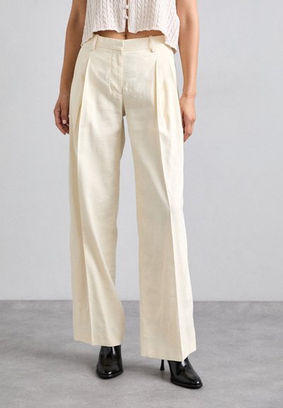 Pantalons larges crèmes avec des plis, fabriqués en tissu léger. Avec une taille cintrée, ils tombent jusqu'à la cheville et sont associés à des talons noirs.