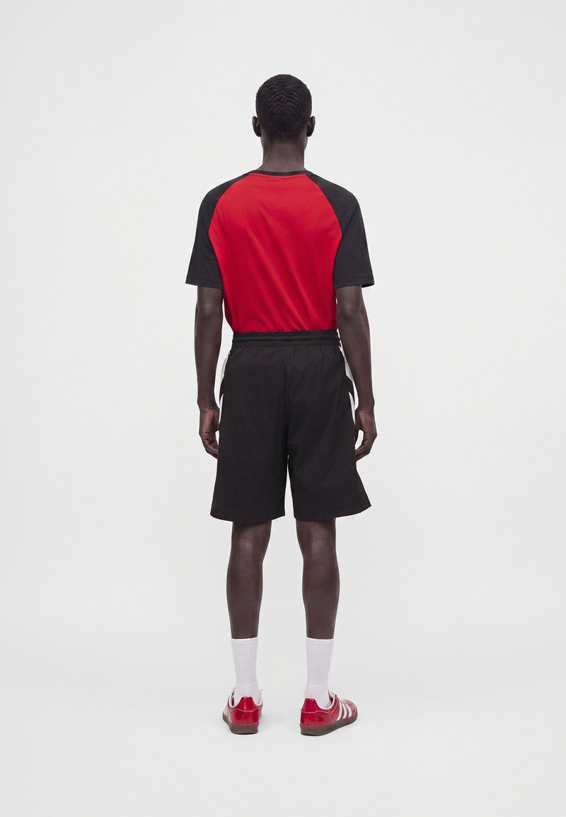Mand iført rød og sort kortærmet skjorte, sorte shorts, hvide sokker og røde sneakers, står med ryggen mod på en ensfarvet hvid baggrund.