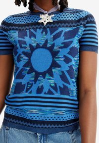 Suéter de punto corto azul con un patrón de explosión solar, diseño a rayas y un intrincado dobladillo de encaje. Tejido texturizado con múltiples tonos de azul.