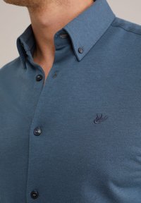 Zila krekliņa ar teksturētu audumu, kuram ir tumši pogas un neliels izšūts logo uz krūškurvja. Tuvplāns pie kakla apļa.