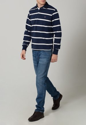 Man walking wearing navy blue and white striped long-sleeve polo, blue jeans, and dark brown leather shoes on a plain background.