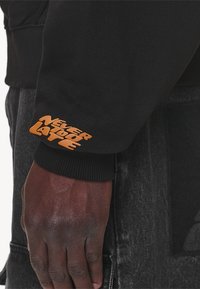 Manica di felpa nera con testo arancione "NON È MAI TARDI." Primo piano di una mano appoggiata su un tessuto scuro, che mette in risalto il contrasto di texture.