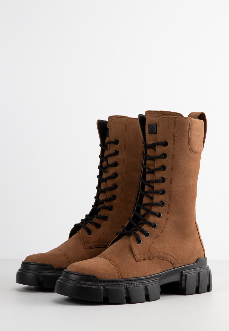 Braune Wildleder-Stiefel im Kampfstil mit schwarzen Schnürsenkeln, verstärktem Zehenbereich, dicker schwarzer Sohle und einer Höhe bis zur Mitte der Wade. Mit einem Logo-Tag an der Seite.