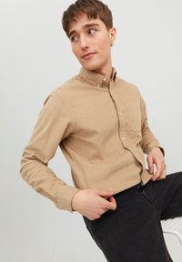 Camisa de pana beige con cuello abotonado, bolsillo en el pecho y mangas largas, combinada con jeans oscuros, sobre un fondo claro.