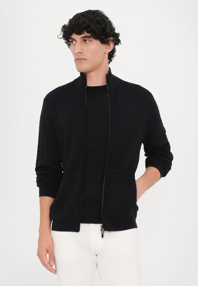 Cardigan negru cu fermoar, cu un model texturat în grilaj, manșete și tiv ribonate, purtat peste un tricou negru cu guler și pantaloni albi.
