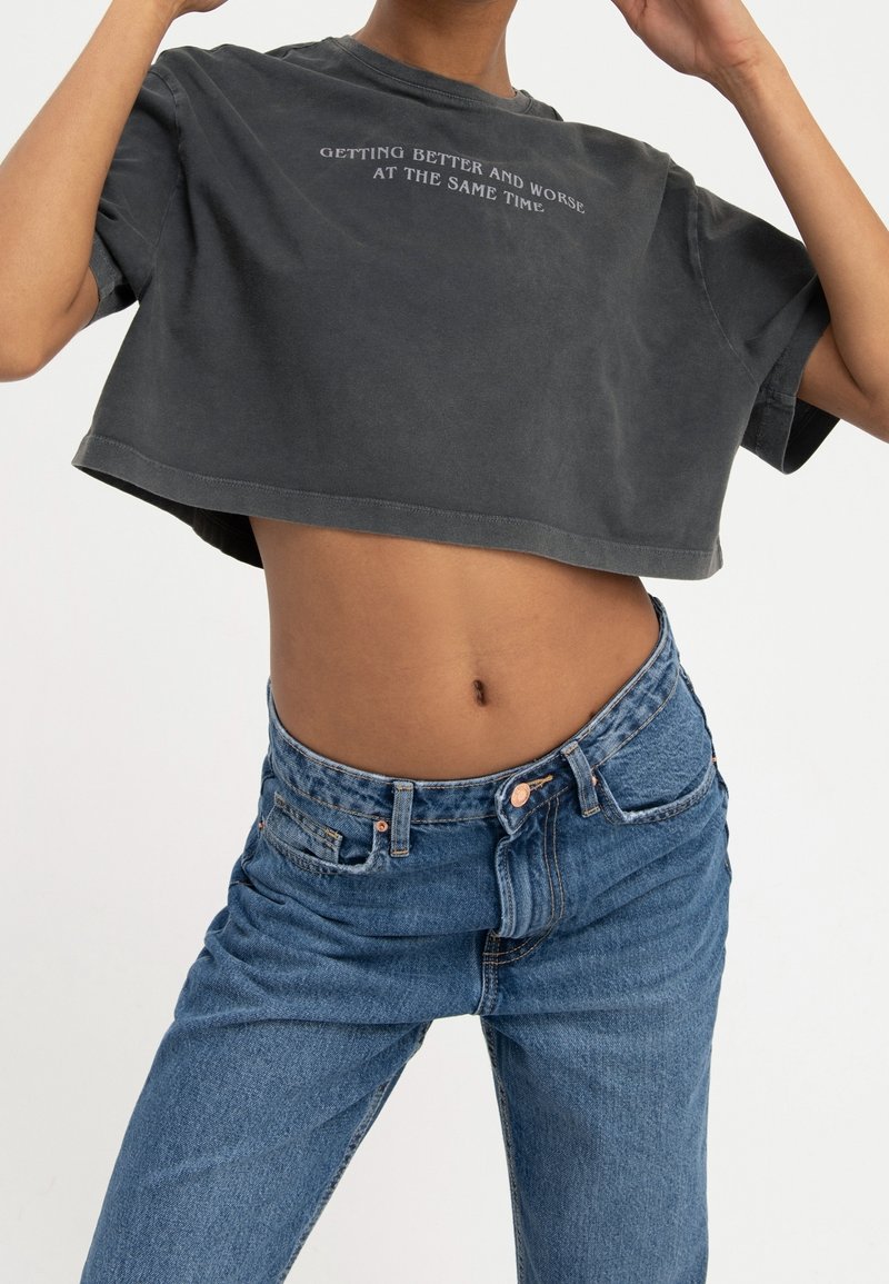 Afskåret mørkegrå t-shirt med tekst trykt på forsiden, parret med højtaljede blå jeans. 100% bomuldsstof, afslappet pasform.