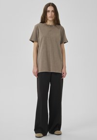 Frau mit langen braunen Haaren, die ein lockeres braunes Kurzarmhemd, schwarze Hosen mit weitem Bein und beige Schuhe trägt, vor einem schlichten grauen Hintergrund steht.