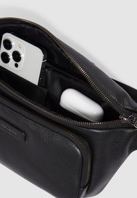 Schwarze Ledertasche mit Reißverschluss. Innen sind ein Smartphone und weiße kabellose Ohrhörer sichtbar. Die Oberfläche zeigt eine strukturierte Optik.