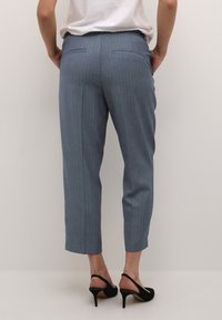 Kvinna iklädd blå randiga cropped byxor och svarta klackiga slingback-skor, som står mot en neutral ljus bakgrund.