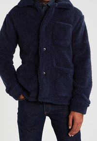 Mężczyzna w granatowej polarowej kurtce z dwoma przednimi kieszeniami i zapięciem na napy, w zestawieniu z ciemnoniebieskimi dżinsami.