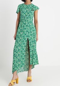 Grön blommig maxiklänning med korta volangärmar, hög sprund och en flödande design. Accenterad med gula klacksandaler.