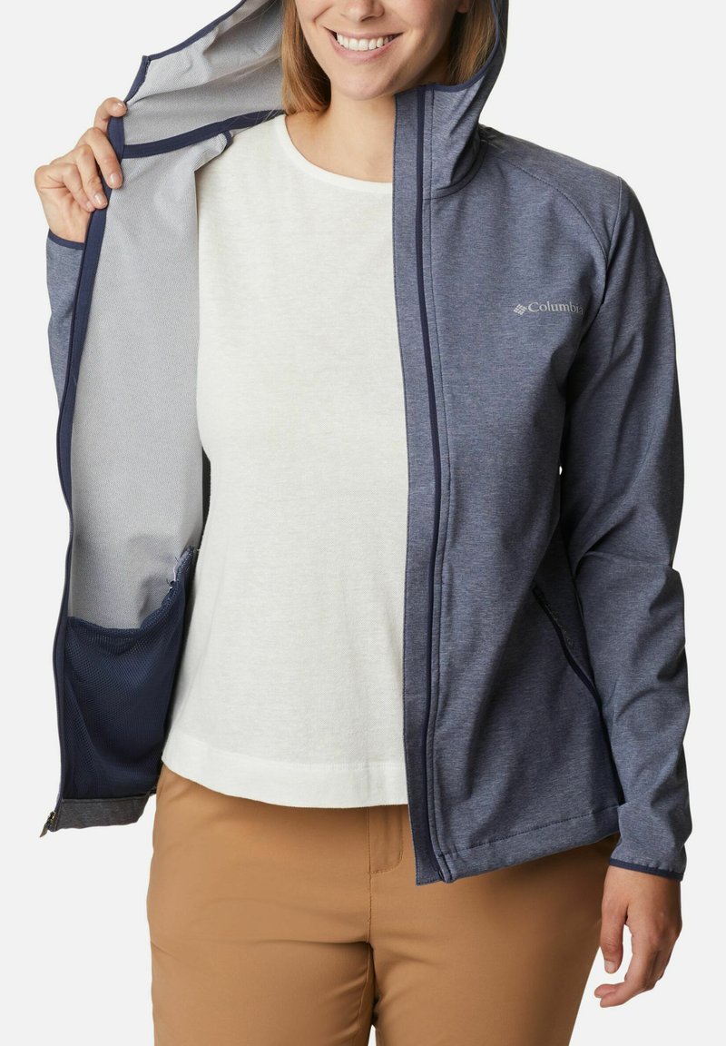 Giacca leggera blu navy con cappuccio, dotata di chiusura con zip, texture morbida e tasca interna in rete. Indossata su una maglietta chiara.