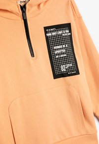 Sudadera color naranja claro con bolsillo frontal, cierre de media cremallera y una etiqueta gráfica negra con patrón de cuadrícula y detalles en texto. Textura suave.