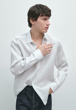 Junger Mann mit dunklem Haar, der ein weißes Hemd mit Nadelstreifen und dunkle Jeans trägt, schaut zur Seite und nimmt mit einer Hand seinen Kragen zurecht.