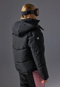 Geacă pufă neagră cu glugă, cu secțiuni orizontale matlasate și un logo pe mânecă. Poartă ochelari de schi și pantaloni roz.