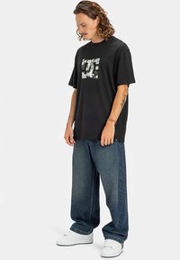 Homem com cabelo encaracolado, vestindo uma t-shirt gráfica preta, jeans azuis largos e ténis brancos, a pé e a olhar para baixo.