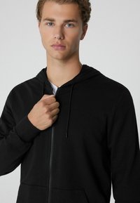 Schwarze Zip-Up-Hoodie mit weicher Textur, Kordelzugkapuze und Vordertaschen. Das Modell hält den Reißverschluss mit einer Hand.