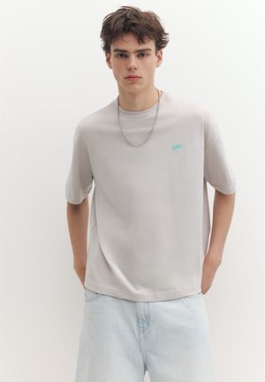 Ung mand med kort brunt hår iført en lysegrå t-shirt og lyseblå jeans står foran en hvid baggrund.