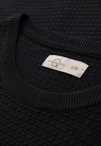 Maglione nero a trama texturizzata con collo a girocollo a costine e un'etichetta che riporta "Quintessence" di taglia M. Il tessuto presenta un motivo intrecciato.