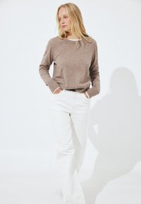 Femme blonde debout, les mains dans les poches, portant un pull beige et un pantalon blanc taille haute, sur un fond blanc.