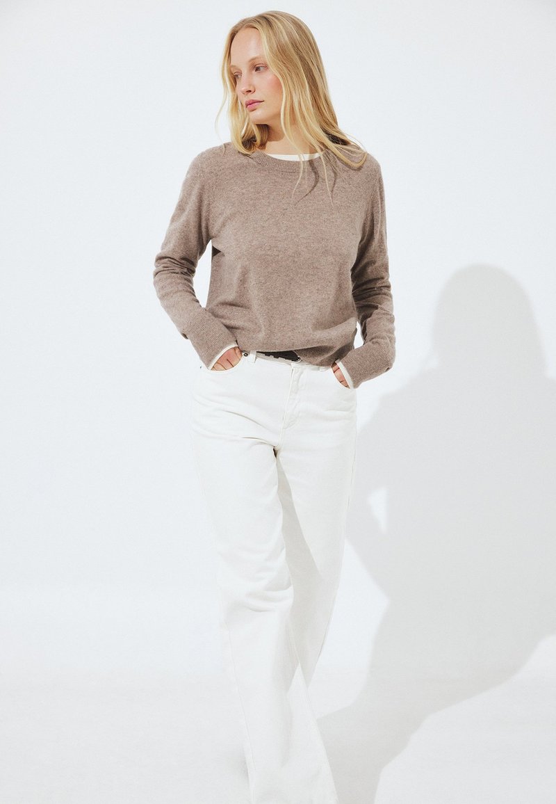 Femme blonde debout, les mains dans les poches, portant un pull beige et un pantalon blanc taille haute, sur un fond blanc.