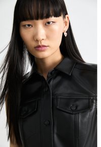 Camicia in pelle nera con colletto a punta, chiusura frontale con bottoni e due tasche sul petto. Il modello ha i capelli neri lisci e orecchini.