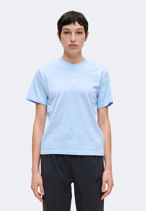 Jeune personne aux cheveux courts et foncés portant un t-shirt Adidas bleu clair et un pantalon Adidas noir sur un fond uni.