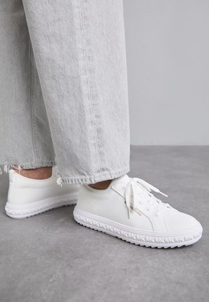 Baskets décontractées blanches à lacets portées avec un jean gris clair raccourci, vues de profil sur un sol gris contre un fond uni.