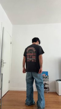 Osoba oblečená v černém vintage tričku Levi's a volných modrých džínách stojí na dřevěné podlaze v minimalistické místnosti.