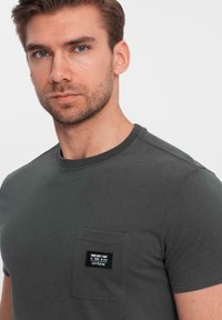 Tmavě šedé bavlněné tričko s krátkým rukávem, kulatým výstřihem a náprsní kapsou na levé straně s visačkou s bílým textem.