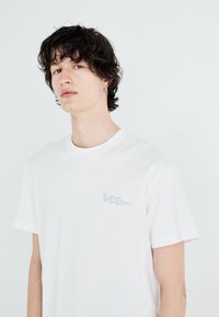 Weißes Baumwoll-T-Shirt mit kurzen Ärmeln, versehen mit einem hellblauen "Lee"-Logo auf der linken Brustseite. Glatte Textur, klassischer Rundhalsausschnitt.
