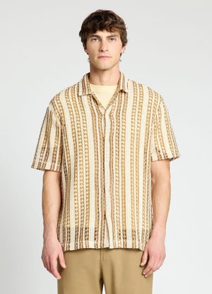 Giovane uomo con capelli castani corti indossa una maglietta a maniche corte lavorata a maglia a righe beige e marroni e pantaloni marrone chiaro su uno sfondo semplice.