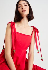 Woman wearing a sleeveless red dress with tied shoulder straps adorned with small red and green beaded tassels, against a plain background.