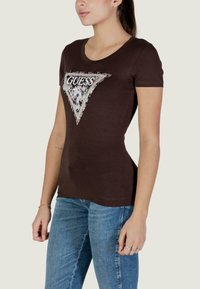 Camiseta ajustada marrón con un logo triangular de estampado animal, adornado con pedrería y la palabra "GUESS" en el centro.