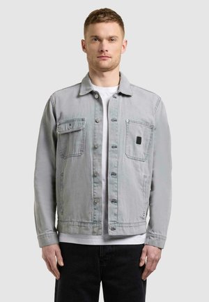 Mann iført lysegrå denimjakke med knapper og brystlommer over hvit skjorte, stående mot en ensfarget lys bakgrunn.