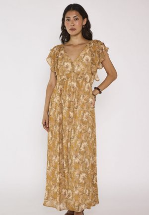 Femme debout portant une robe longue jaune à imprimé fleuri, manches courtes à volants et col en V, posant devant un fond uni clair.