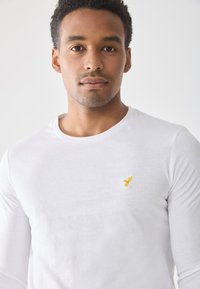Camiseta blanca de manga larga con cuello redondo, que presenta un pequeño bordado de un pájaro amarillo en la parte izquierda del pecho. Textura de algodón suave.