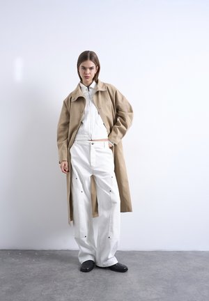 Frau steht vor einer weißen Wand und trägt einen beigen langen Mantel, eine weiße Jacke, weite weiße Hosen und schwarze Schuhe, mit einer Hand in der Tasche.