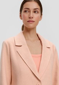Jasnoróżowa marynarka z wełny z wklęsłym kołnierzem, z pojedynczym zapięciem na guzik i miękką fakturą, noszona na dopasowanej bluzce z prążkowanej tkaniny.