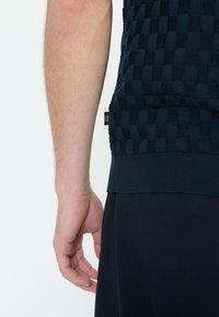 Moška roka visi ob torzu, oblečena v temno modro teksturirano srajco s kratkimi rokavi in temno modre hlače na belem ozadju.