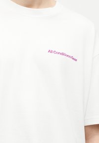 Λευκό μπλουζάκι με μικρό ροζ κεντημένο κείμενο «All Conditions Gear» στην αριστερή πλευρά του στήθους, που φοριέται από έναν άνθρωπο που κόβεται στο λαιμό και το χέρι.