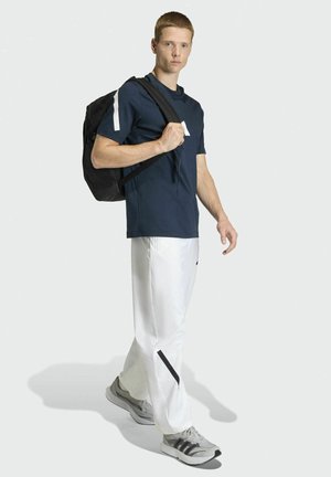Young man walking wearing a navy shirt, white pants with black accents, gray sneakers, and carrying a black backpack over one shoulder.