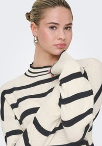 Ivoorkleurige geribbelde trui met zwarte horizontale strepen, hoge kraag en lange mouwen. Voorzien van duimgaten bij de manchetten voor extra comfort.