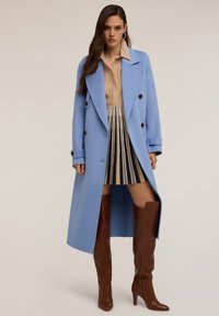 Jasnoniebieski wełniany płaszcz o podwójnym zapięciu, z szerokimi klapami i czarnymi guzikami, w połączeniu z beżową koszulą i plisowaną spódnicą w paski.